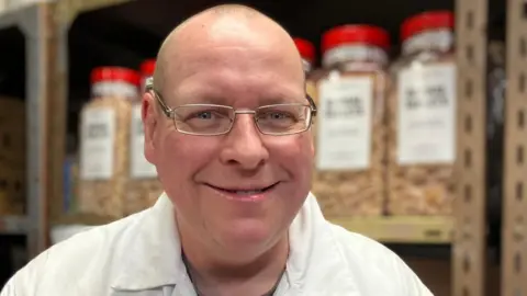 A man with glasses is standing in a white coat smiling and looking at the camera. Behind him are big see through jars full of orange hard boiled sweets. 