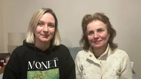 Two women sit side by side indoors on a sofa; one wearing a black hoodie with a Monet print, the other in a light-coloured knitted cardigan. A lamp and small table are visible in the background.