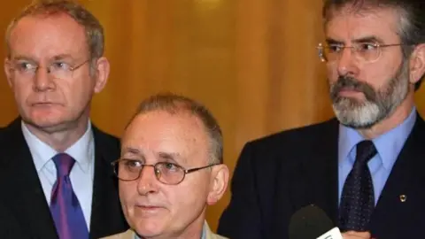 PA Media Martin McGuinness, Denis Donaldson and Gerry Adams are all looking to their right. All three are wearing glasses. Mr McGuinness and Mr Adams are both wearing dark suits, shirts and ties. Mr Donaldson is wearing a tanned coloured jacket and a denim shirt. There are three microphones in front of them.
