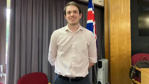 A man with short brown hair is smiling at the camera. He is wearing a pale shirt tucked into dark trousers and has his hands behind his back.  Behind him are grey curtains and a Union Flag.