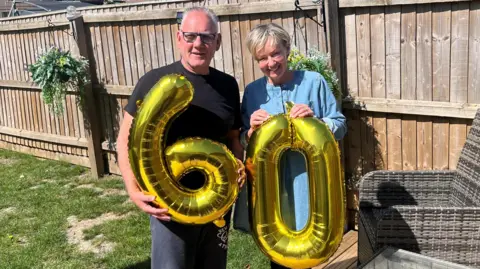 Tony Raine has short spikey grey hair with dark framed glasses. He has a black t-shirt on with grey jogging bottoms and is holding an inflatable number six in gold colour. Tracy Raine is holding a similar number 0 and has a denim dress with short blonde hair. Behind them is a garden fence with some grey rattan furniture on the right.