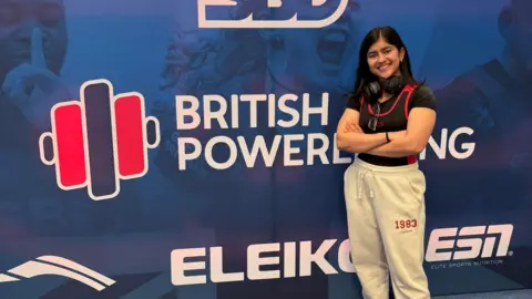 A girl standing with her arms folded smiling in front of a sign which says British Powerlifting