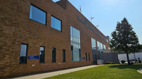 Exterior view of a hospital building. It has brown bricks and a row of tall oblong windows. In the foreground is a pavement, a tree and a grassy area. People can be seen in the distance.