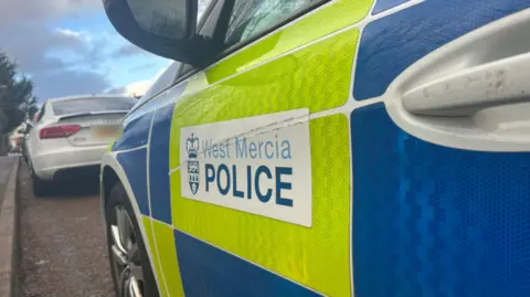 A close up image of a police vehicle's door. The door is predominately white with yellow and blue high viz stickers on the side as well as a white sticker that reads "West Mercia Police". 