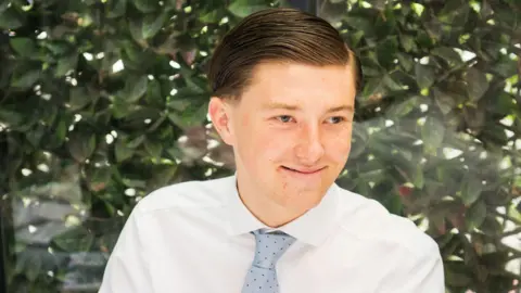 A young man smiling wearing a white shirt and a blue tie. He is sitting in front of a green bush. He has brown hair.