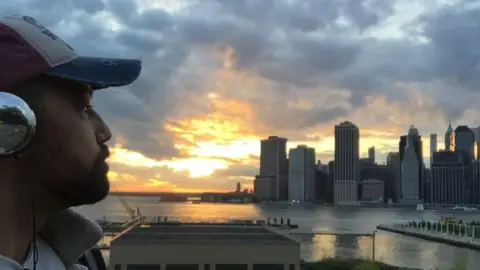 Stephen Hart Side-profile view of a man wearing headphones and a baseball cap, looking across a river toward a city skyline at sunset.