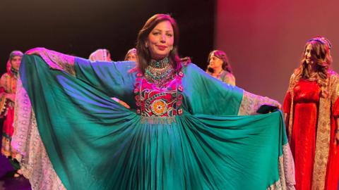A woman wears a flowing green dress with colourful embroidery
