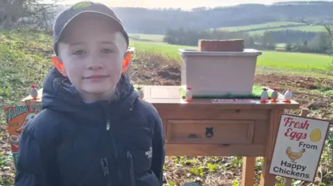 A young boy wears a coat and cap. He is smiling at the camera while standing in front of a wooden desk with signs reading "Sam's Eggs". On the table are two grey plastic boxes, both with a brick on top, and small chick-themed toys.