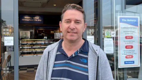 Joe Pinzari stands in front of the entrance to The Bakery with glass doors. The shop interior shows display counters with baked goods and signs. Joe's wearing a striped shirt and a light grey hoodie. Signs with prices and information are visible on the windows beside the entrance.