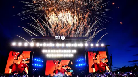 BBC/Sarah Jeynes/Jamie Simonds Fireworks explode above a stage branded "BBC Radio 1" as Chris Martin, glowing in orange light, smiles on three large screens around the stage