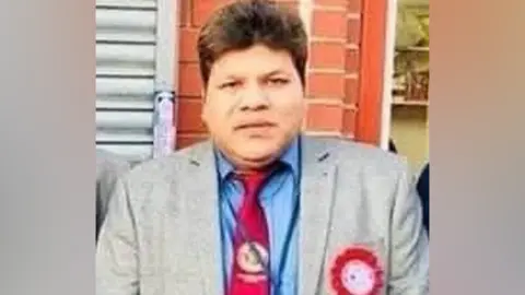 Other A man with brown hair and wearing a grey jacket, blue shirt, red tie and a red rosette.