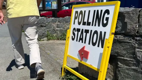 The photo shows a polling station sign with a red arrow on it. A man in beige trousers is walking by.