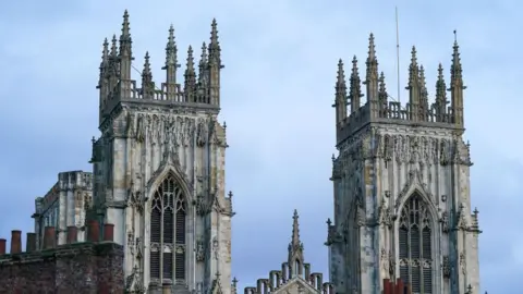 Two tall Gothic cathedral towers rise into the sky, detailed with intricate stone carvings and pointed spires.

Large arched windows and decorative statues cover the brickwork, showing traditional medieval architecture.

The cloudy sky and weathered stone give the structure a historic and grand appearance.