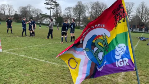 A number of players from the Brighton and Hove Sea Serpents rugby club on the pitch at Hove recreation ground. The players are all wearing navy kits and there is a club flag in the foreground of the image.