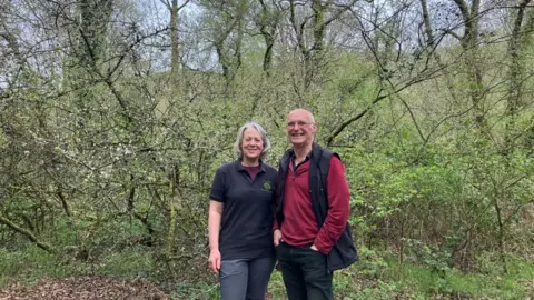 Michelle Grist and her partner, Kevin Chamberlain who are co-owners of Wray Valley Camping. They are standing together in a woodland clearing. Michelle is wearing dark grey trousers and a black polo top, she has white hair and is smiling. Kevin is wearing black trousers, a red fleece top and a black gilet. He is wearing glasses and smiling.