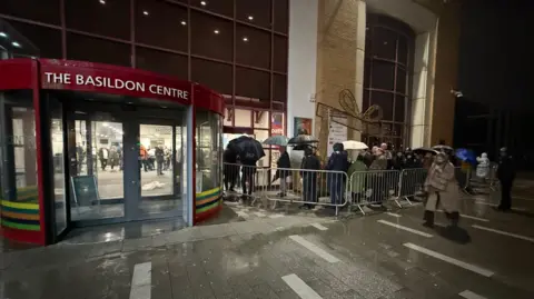 BBC/Simon Dedman A building with a revolving door painted red saying the basildon centre. To it's right in the centre of the frame is a queue of people many holding umbrellas and entering a side door of the council building.