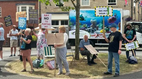 Sally Howard A group of people with placards stand on a green area to protest a warehouse development.