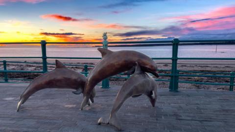 Bronze statue of three dolphins leaping by the seafront with a background of a vivid blue, pink and orange sky.