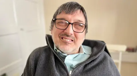 David Cording is pictured sitting down in a room with pale walls. He is wearing a dark grey fleece with a zip over a light blue shirt. He has glasses, light stubble and short dark hair and he is smiling. His eyes are almost closed.