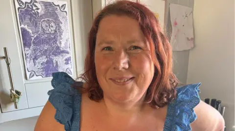 Abbie Barton has shoulder-length red hair and is wearing a blue sleeveless top. She is standing indoors in front of a framed owl print and children's artwork. She is smiling.