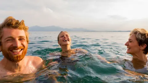 Getty Images Cheerful young woman splashing water while enjoying swim with friends in sea on sunny day