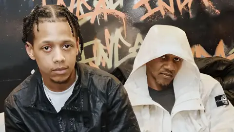 ML and Giggs sit on a sofa, ML leaning forward in front of his dad. ML wears a dark short sleeved shirt while Giggs wears a long sleeved white jacket with the hood up. They're pictured in front of a dark wall that's been graffitied. 