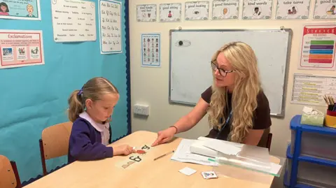 BBC/Kate McGough A blonde-haired little girl wearing purple school dress and cardigan sits at a table with a woman with long blonde hair and glasses wearing a brown top. The woman is point at a phonics card in front of the little girl with a picture of a strawberry on it. They are in the corner of a classroom with displays of words on the wall.