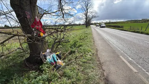 Richard Knights/BBC A tree on the side of a road. By the tree there are flowers and a red football shirt 