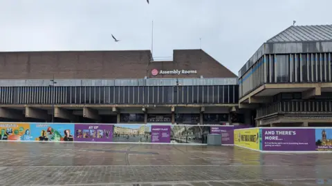 The Derby Assembly Rooms building surrounded by walls which have advertising displaying developments in Derby
