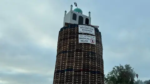 An effigy of a mosque is on top of a bonfire built out of wooden pallets. Signs say secure our vorder and end the threat of radical Islam. The sky is cloudy. 