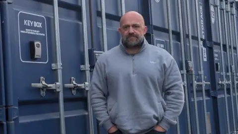 A bald man with a stern expression looking towards the camera, he's wearing a light blue jumper and black jeans. He stands in front of blue storage containers.