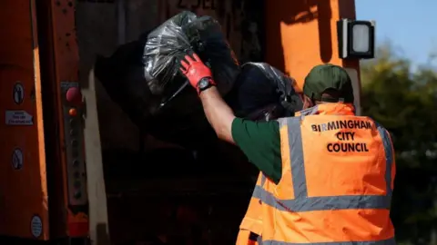 Reuters A man wearing a green hat, green shirt and an orange hi-vis jacket has his back to us as he throws two, full black bin bags into the back of a refuse vehicle.