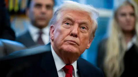 Donald Trump appears in a dark suit and red tie in the Oval Office