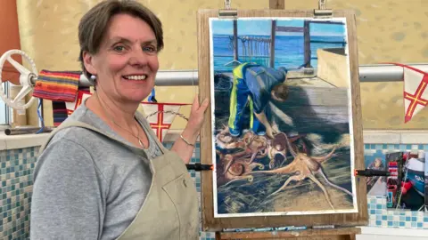 BBC News A woman wearing an apron and smiling. She is next to a pastel painting of a man who is leaning over some octopus which he just caught