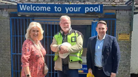LDRS Three people are standing in front of a sign which says Welcome to your pool. On the right is MP Sam Rushworth who is about 50 and wearing a blue jacket and light blue shirt. The man in the middle is older with glasses and a white beard and wearing a hi-vis jacket and holding a helmet. The woman on the left is middle-aged, blonde and wearing a pink dress 