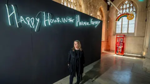 Richard Walker/ImageNorth A woman with mousy brown shoulder-length hair standing in a full-black outfit in front of a black billboard with white neon lettering spelling out "Happy Hour in the Harmful Factory".