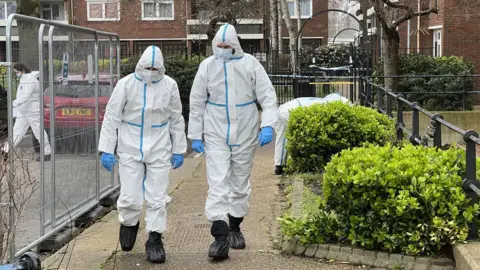 Steve Hubbard/BBC Forensic officers walk along a path at the crime scene of a home