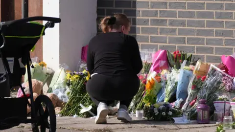 PA Media A woman dressed in black bends down to look at the flowers, with a pushchair nearby