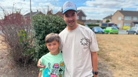 A young boy with brown hair who is smiling at the camera. He is wearing a mint green t-shirt which has a sun and a skeleton on it, and the skeleton is surfing and wearing a pink hat. Next to him is his dad, who has his arm around the boy. The man has dark stubble and is wearing a white and blue cap and a white t-shirt