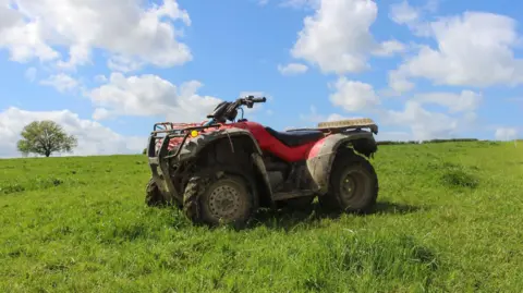 Getty Images A quad bike in a field. It is a sunny day, the sky is blue with white clouds. The quad bike is in the middle of the photo, it is black and red.