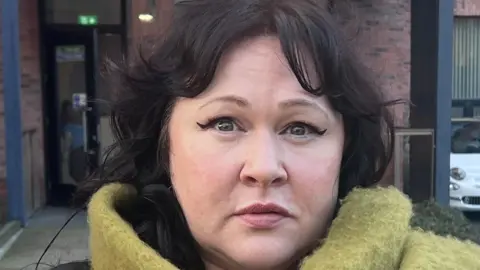 Sara Morrison is wearing a brown top with a large green scarf around her neck. She is standing in front of a red brick building with black windows. There are cars and a motorbike in a car park between her and the building. The path around the building is of white stones. 