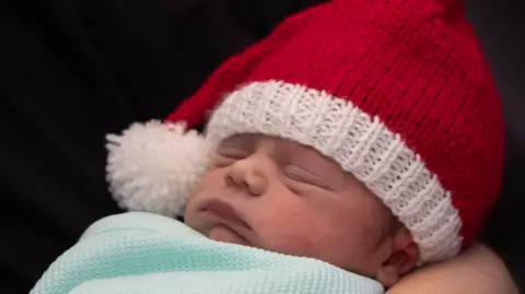 A newborn baby with a santa hat on and swaddled in a light blue blanket. 