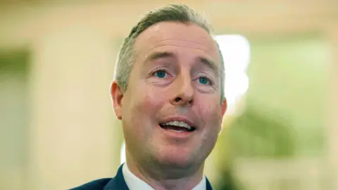 Paul Givan is standing inside the Stormont Building, he is speaking to press. He has short white hair and is wearing a dark suit. 