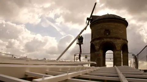 The white windmill sails lay on the ground below the round windmill, before being hoisted into position. There are two people stood on a hoist that is elevated alongside the windmill.