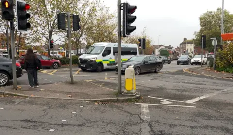 A picture of the junction from Mansfield Road to Hurdle Way.  The picture shows traffic at the junction and a person waiting to cross the traffic lights.