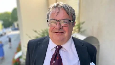 Middle-aged white man with glasses smiling with lilac shirt and burgundy tie and navy blazer 