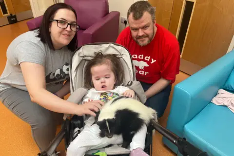A woman with shoulder-length brown hair and a man with brown hair and a beard smile at the camera. Between them is a young girl with brown hair sitting in a pram. She has a black and white skunk on her lap.