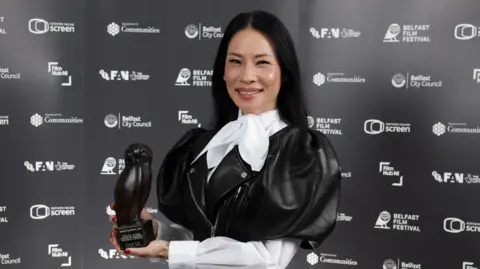 Lucy Liu smiles while posing with her award at the Belfast Film Festival.  She has long, straight, black hair and is wearing a black leather frock coat with puffed sleeves over a white blouse tied with a large bow at her neck.  She is holding an award shaped like an owl. 