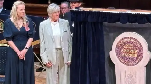 Lissie Harper and Mrs Michael Winner, who has short, white hair and wears a loose, grey suit, stand on a wooden stage next to a stone memorial in Andrew Harper's name. Curtains have been opened on either side to reveal the memorial.