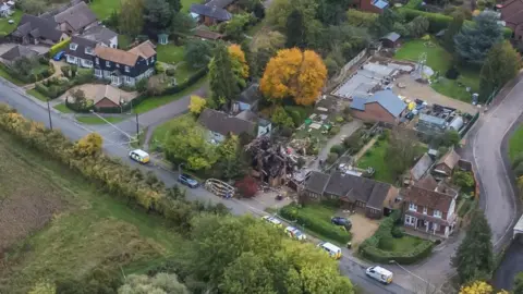 Drone photo shows destroyed buildings with emergency vehicles parked outside. Nearby homes appear undamaged.